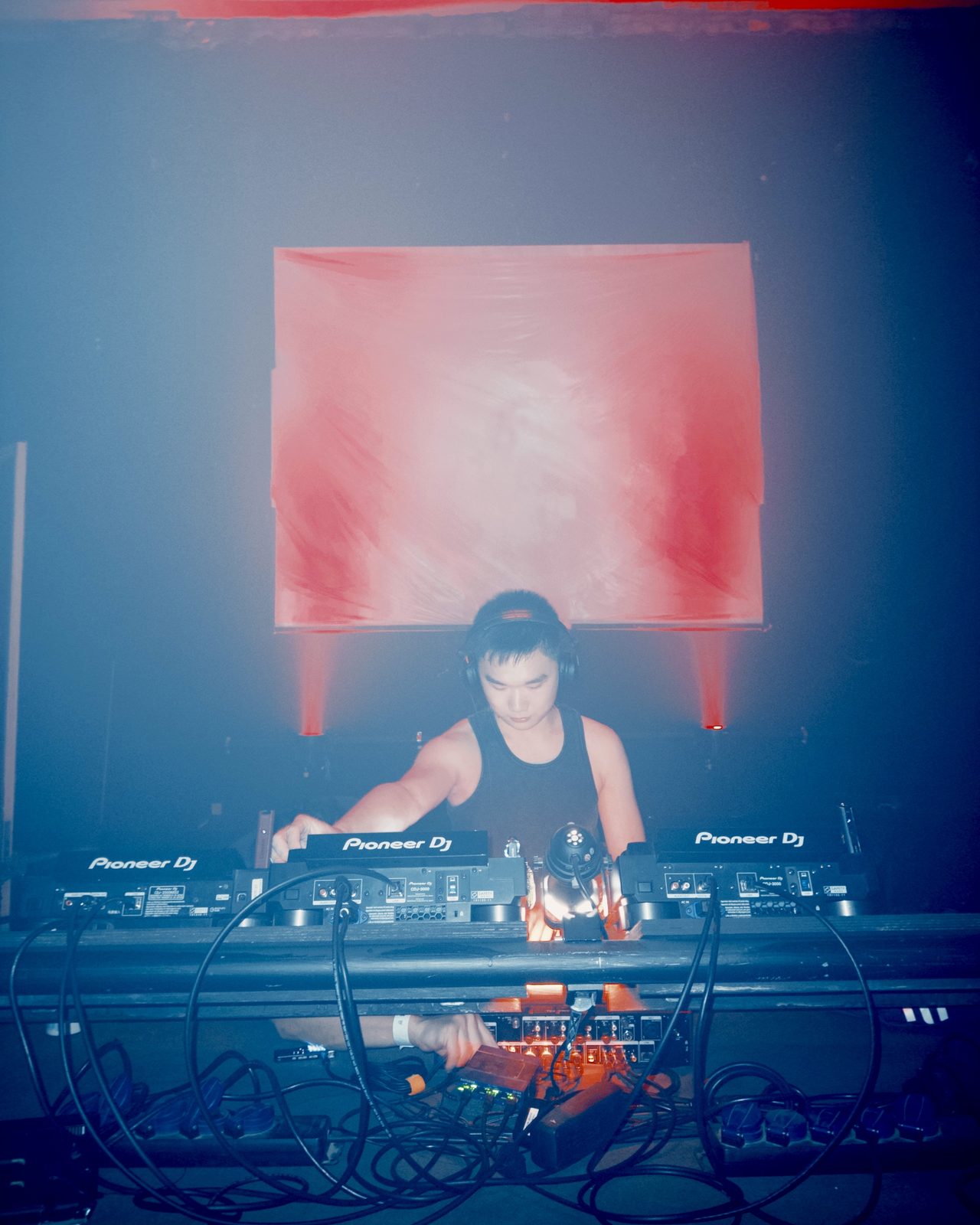 DJ in headphones at three Pioneer decks, a red-lit screen behind
